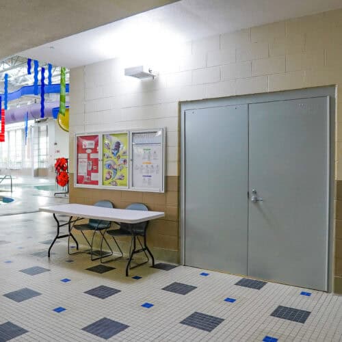 A hallway featuring a closed gray Smooth Pultruded Fiberglass Door, a bulletin board with notices, and a small table with two chairs. Colorful decorations hang in the background, and there is a tiled floor.