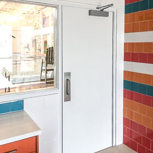 A white Smooth Pultruded Fiberglass Door with a window beside it is partly reflected in a mirror. The wall is tiled with red, white, orange, green, and blue stripes.