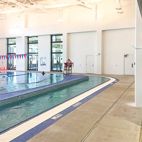Indoor swimming pool area with a lifeguard seated on a chair near the poolside and lanes marked with blue and white tiles, featuring an AF-100 smooth pultruded fiberglass door for easy access.
