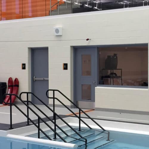 Indoor swimming pool ladder with metal handrails, leading into the water. Two smooth pultruded fiberglass doors and a large window are visible in the background against a white wall. Red safety equipment is on the left.