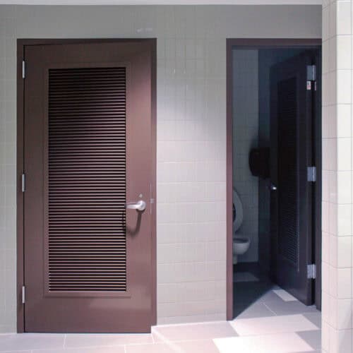 A tiled restroom with two brown, AF-100 pultruded fiberglass stall doors slightly ajar, revealing a toilet in one.