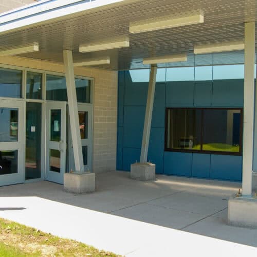 A modern building entrance with large glass doors, blue paneling, and metal support beams showcases an AF-220 Composite Fiberglass Door. There's a window to the right and a concrete walkway featuring a subtle sandstone texture leading up to the doors.