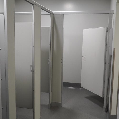 A public restroom with multiple stalls featuring commercial bathroom partitions. Some of the stall doors are open, revealing neatly partitioned toilet areas. The floor is covered with gray tiles.