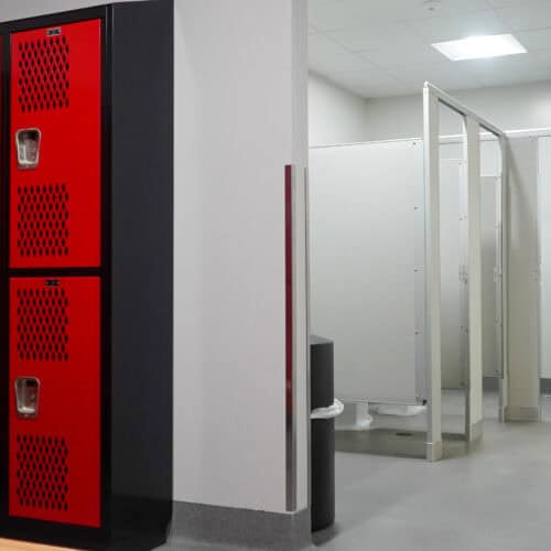 A gym locker room with red lockers on the left and a row of bathroom stalls fitted with commercial bathroom partitions on the right, separated by a wall. The floor is light gray, and the walls are mostly white.