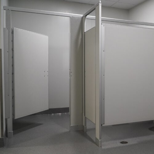 A public restroom with two stalls, one with an open door and the other partially closed, both featuring light grey commercial bathroom partitions and clean, minimal decor.