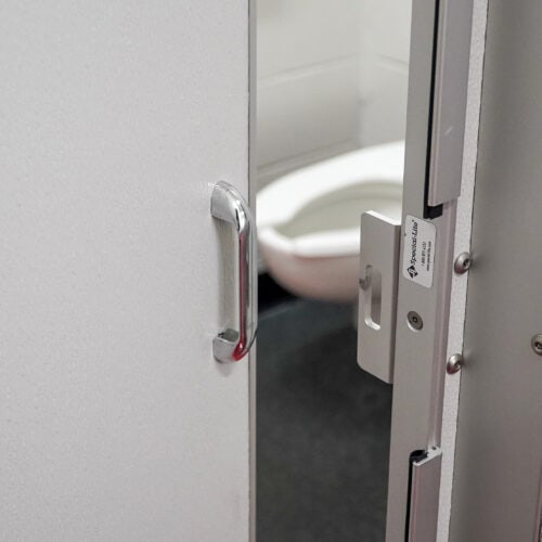A partially open restroom stall door reveals a white toilet inside. The door, part of the commercial bathroom partitions, features a metal handle and a hinge lock mechanism labeled 