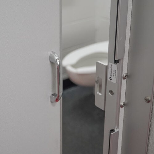 A partially open bathroom door reveals a white toilet inside, framed by commercial bathroom partitions. The door features a metal handle and a commercial lock.