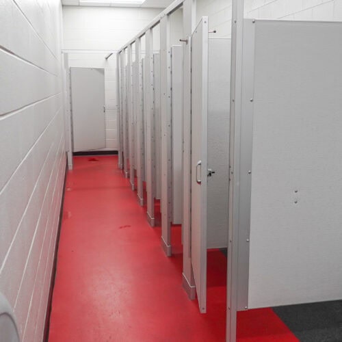 A row of white bathroom stalls with commercial bathroom partitions on the right side of a narrow hallway featuring red flooring and white walls. An open door is visible at the end of the hallway.