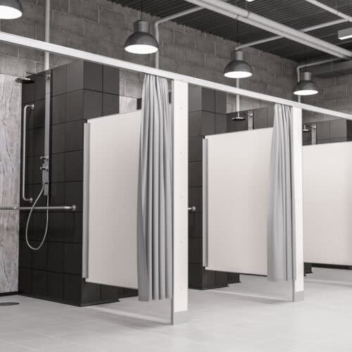 The interior of a modern public shower facility features dark tiled walls and elegant commercial bathroom partitions as white dividers, complemented by gray curtains. Ceiling lights illuminate the space, highlighting its clean and minimalist design.