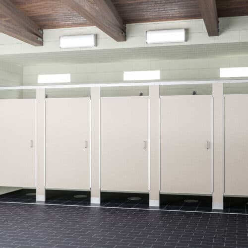 A row of four beige commercial bathroom partitions with closed doors stands under the glow of overhead fluorescent lighting. The floor is covered with dark tiles.