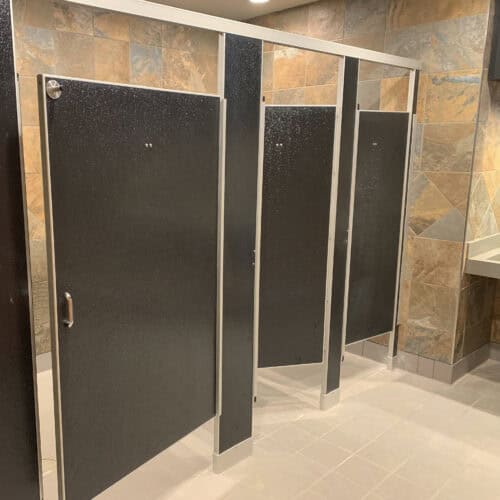 A public restroom with three black metal commercial bathroom partitions and tan tiled walls, featuring a sink countertop in the background.