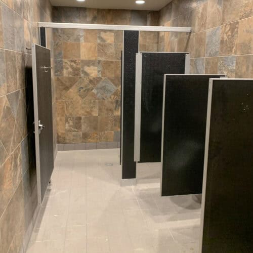 A clean, tiled public restroom with several black commercial bathroom partitions. The floor is light gray and the walls have a mix of brown and gray tiles. Ceiling lights illuminate the area.