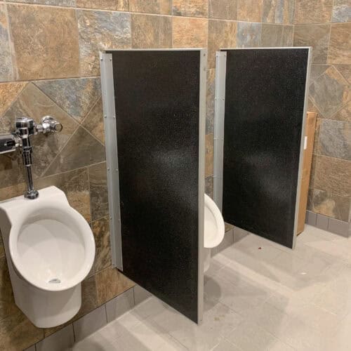 A restroom with two urinals separated by black commercial bathroom partitions, set against a wall with stone tile.