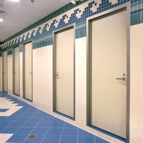 Row of four closed restroom doors in a tiled public facility, featuring blue and white wall and floor tiles with a geometric design near the ceiling, showcasing modern commercial bathroom partitions.