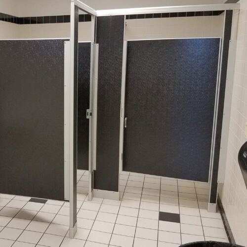 A restroom with two black commercial bathroom partitions, closed doors, and beige tiled walls and floor.