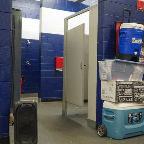 A locker room with blue and white walls. A shower and commercial bathroom partitions are visible on the left. Stacked plastic bins and coolers are on the right. A black speaker is on the floor near the entrance.