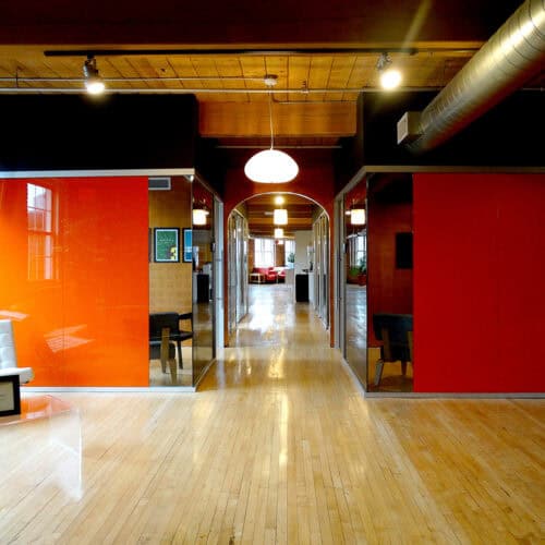 The interior corridor, featuring orange walls and glass partitions with aluminum framing, leads to a brightly lit space at the end. The wooden floor adds warmth, while round ceiling lights above complete the modern design of this LiteSpace.