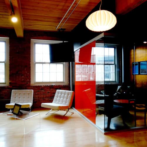 Modern office space with two white lounge chairs, a glass table, brick walls, and large windows. A hanging lamp and a partitioned meeting area with a red wall add character while sleek aluminum framing throughout creates a refined LiteSpace interior.