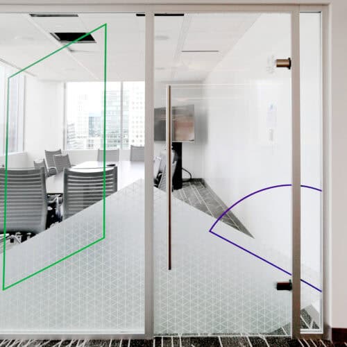 A modern office meeting room, featuring LiteSpace frosted glass doors with geometric designs in green and purple, stands out with several grey chairs and a round table inside. The cityscape is visible through the windows, while sleek aluminum framing adds a contemporary touch to the interior.
