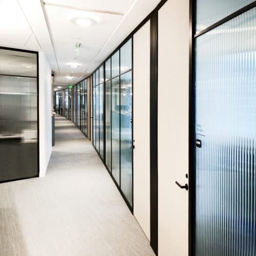 A modern office corridor with LiteSpace frosted glass walls and doors on the right side, featuring a curved path lined with overhead lights, neutral-colored carpeting, and sleek interior aluminum framing.