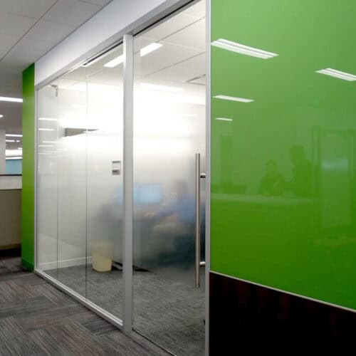 A modern office with green walls and frosted glass doors and windows, framed by sleek Aluminum Framing, showing vague silhouettes of people inside a conference room.