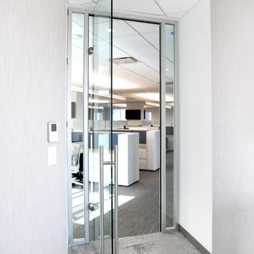 A modern office entrance with a sleek glass door and aluminum framing leads to a brightly lit workspace with white desks and partitions, epitomizing LiteSpace design.