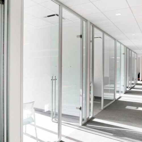 A bright, modern office hallway with glass-walled offices on both sides and natural light streaming in through the LiteSpace aluminum framing. A chair is partially visible in one of the rooms.