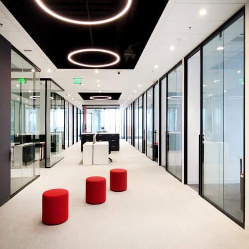 A modern office hallway with glass-walled rooms featuring Aluminum Framing, red stools in the center, black desks, and circular ceiling lights from LiteSpace.