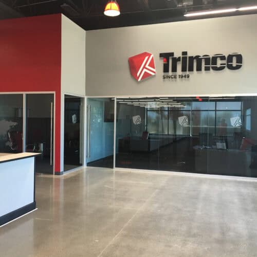 The reception area of a modern office showcases large glass-walled conference rooms with sleek aluminum framing. The wall features the Trimco company logo and slogan 