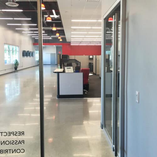 A modern, well-lit office space from LiteSpace featuring a glass entrance with aluminum framing, open work areas, and a row of computers. The red and white color scheme is complemented by ceiling lights and a plant in the corner, creating an inviting interior.