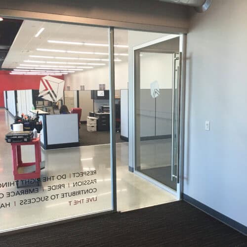 A modern office space with aluminum framing and glass doors, featuring cubicles and wall text about respect, passion, and success. It boasts an industrial design theme with red and white accents, exemplifying the sleek LiteSpace interior.