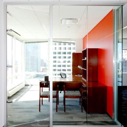 A modern office with glass walls framed by sleek Aluminum Framing, a wooden desk, two chairs, and a red accent wall. Large windows provide a view of tall buildings outside, perfect for the LiteSpace interior.