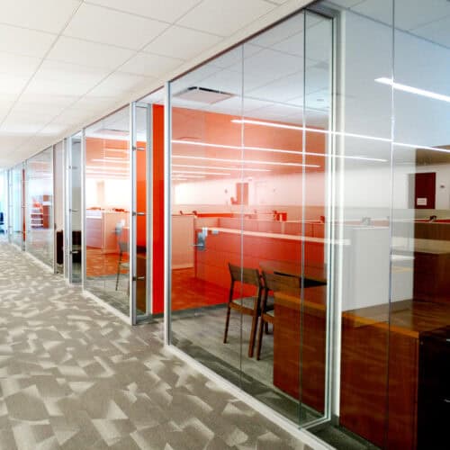 Modern office space with glass partitions by LiteSpace, featuring a red and white color scheme and patterned carpeting. Several desks and chairs are visible through the glass walls, complemented by sleek interior aluminum framing.