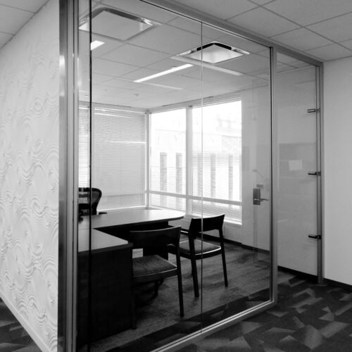 A small, empty office with glass walls framed in sleek aluminum, a desk, chair, and overhead lighting. White patterned wall on one side and large windows allowing in natural light, creating an inviting interior perfect for any LiteSpace setup.