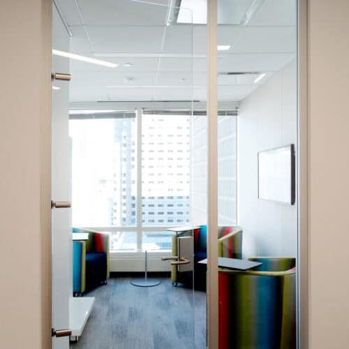 A modern LiteSpace office room with glass walls, aluminum framing, two colorful chairs, a small table, and a large window showcasing a cityscape. The room features a minimalist design with a wall-mounted screen.