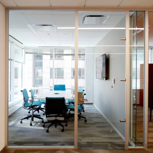 Modern LiteSpace conference room with glass walls, a table, office chairs, a large TV monitor on one wall, and large windows letting in natural light. Framing the sleek design are Interior Aluminum features that add a contemporary touch.