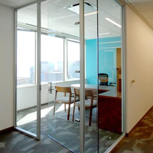 A modern conference room with glass walls framed in sleek aluminum, featuring a wooden table and three chairs. The well-lit space, by LiteSpace, offers large windows with an impressive view of the cityscape.