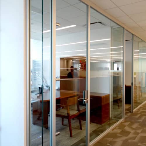 The LiteSpace glass-walled office features a wooden desk, a chair, and aluminum framing partitions separating workspaces. A person is visible inside, while another stands near office equipment. Ceiling lights illuminate the interior.