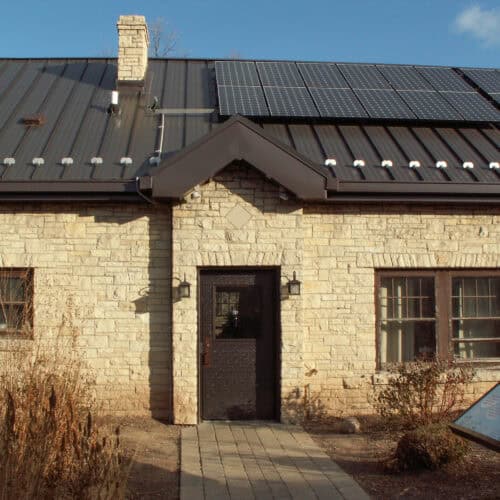 A stone house with a dark metal roof featuring several solar panels. The front door, a Hybrid FRP Door, is flanked by two windows, and a brick path leads up to the entrance. Bushes and a small sign are visible in the yard.