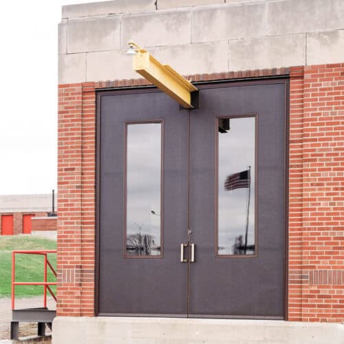 Double doors with tall windows on a brick building, a yellow beam extending above them, and an American flag reflected in the glass. The SL-17 Hybrid FRP Door features an attractive pebble grain finish that seamlessly blends durability with style.