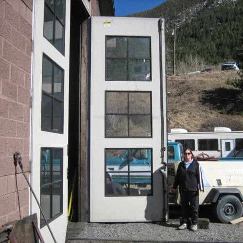 A person stands beside a very tall open Hybrid FRP Door of a building. In the background, a mountain and several vehicles are visible.