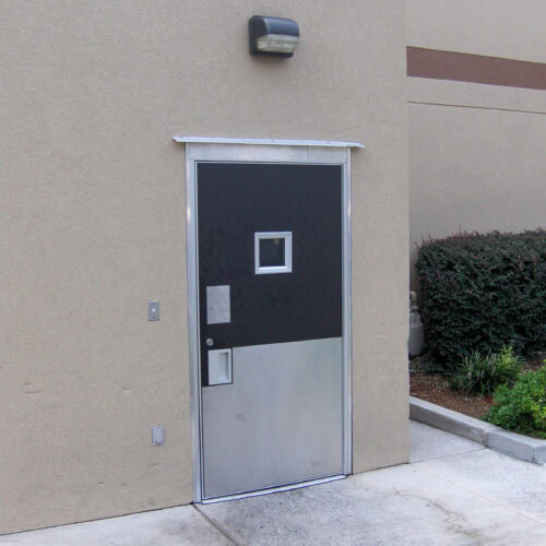 A Metal FRP Door with a small window and mail slot, set in a beige wall under an exterior light fixture. Shrubs and a small garden are visible to the right.