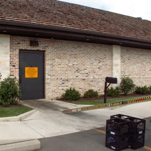 A building with a brick exterior features an SL-17 FRP Door adorned with a yellow warning sign. A mailbox sits beside a barrier while several black crates are stacked on the ground in front.