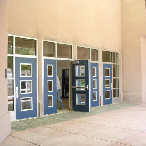 Blue double Pebble Grain Hybrid FRP Doors with window panels, part of a larger building facade, with one door partly open and a person partially visible inside. The exterior has a beige stucco finish.
