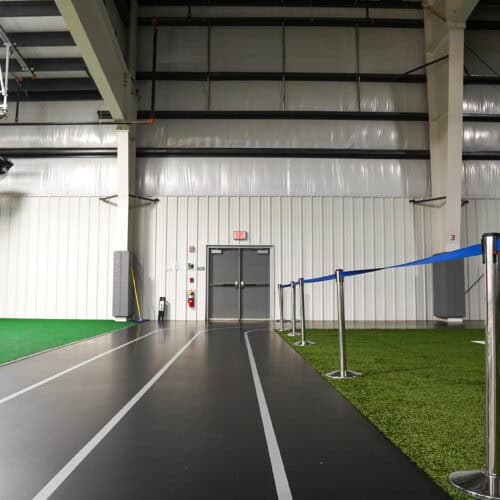 Indoor track and field area with a black running track and adjacent green artificial turf, separated by blue stanchions. Double doors featuring Hybrid FRP Door technology are located at the back of the room.