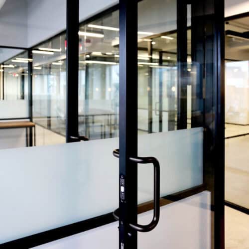 Close-up of a modern glass door with a black metal frame and handle, partially open, leading into an office space with interior aluminum framed glass doors and visible interior lighting.