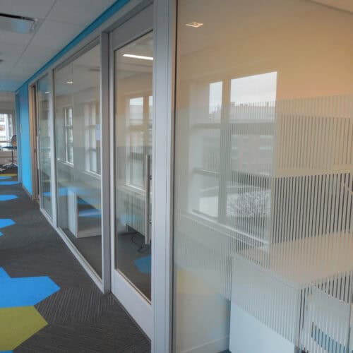 A modern office hallway with glass-walled rooms featuring sleek aluminum-framed glass doors on the right side. The floor boasts a geometric pattern in blue and gray, while windows allow natural light to flood the space.