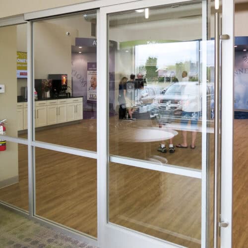 Interior aluminum-framed glass doors lead to a reception area with a wooden floor. Through the glass, a few people are seen standing and one person walking outside in a parking lot.
