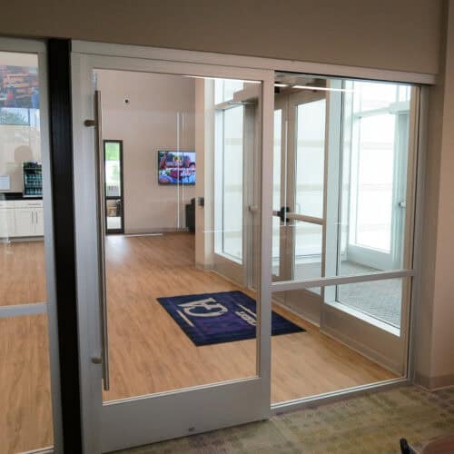 Interior aluminum-framed glass doors open to a lobby with wooden flooring, a mat featuring the logo, and a screen displaying images on the wall in the background.