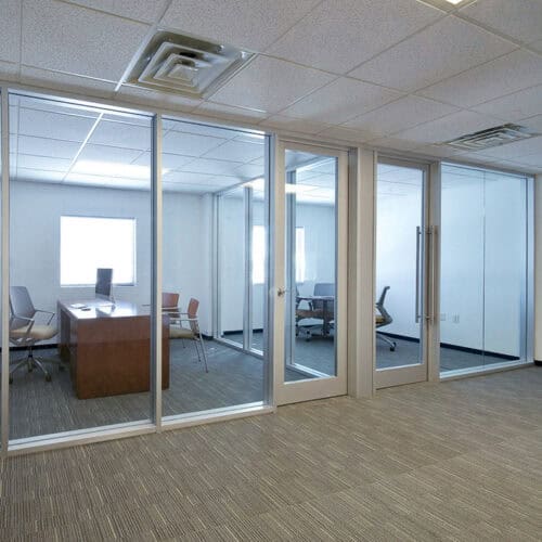 A modern office space with aluminum framed glass-walled rooms featuring desks, chairs, and a carpeted floor. The ceiling has recessed lights and air vents.